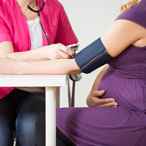 Pregnant woman seated at a table while a healthcare provider fits her with a blood pressure cuff.