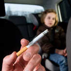 A hand holding a lit cigarette is visible inside a car with a young girl sitting in the backseat. 