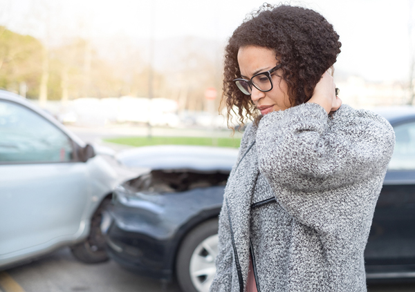 Woman standing in front of a two car crash, massaging the back of her neck, grimacing in pain.