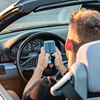 Teenage boy uses cell phone with two hands behind the driver’s wheel.