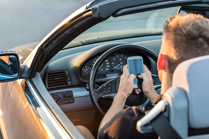 Teenage boy uses cell phone with two hands behind the driver’s wheel.