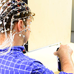 Doug is wearing the fNIRS device, which looks like a fishnet cap with sensors and wires. He is sitting across from Helga, and there is an object, a coffee thermos, between them on the table.