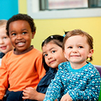 Five toddlers are standing or sitting in a row, smiling. The group is ethnically diverse and a mix of boys and girls.