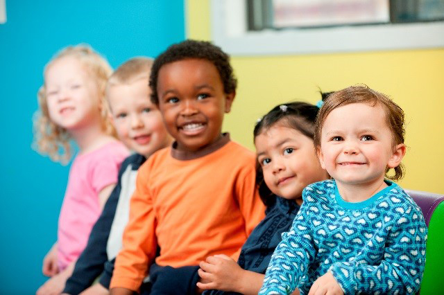 Five toddlers are standing or sitting in a row, smiling. The group is ethnically diverse and a mix of boys and girls.