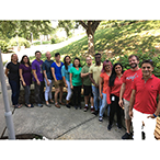 A group of researchers stand in a curved line, arranged like a rainbow according to the color of their shirts.