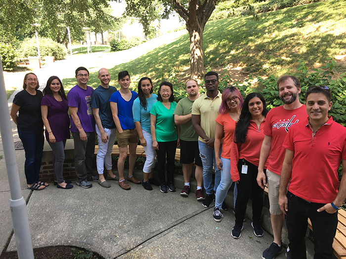 A group of researchers stand in a curved line, arranged like a rainbow according to the color of their shirts.