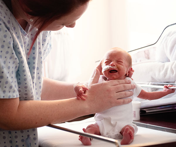 Woman in a hospital gown helping a premature baby sit up in an infant bed.