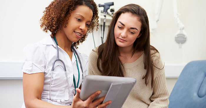 A young woman in a healthcare setting is looking at a digital tablet with her healthcare provider.