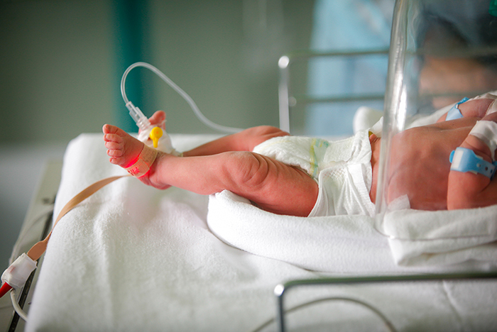 An infant in an incubator, with an oxygen sensor on one foot and an intravenous line on the other foot.