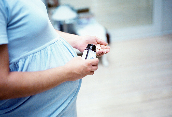 Side view of the torso of a pregnant woman holding a medicine bottle in one hand and pills in the other.