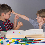 Two boys seated at table, one with fists clenched the other with hand on first child’s forehead.