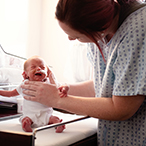 Woman in hospital gown supports preterm infant seated on table.