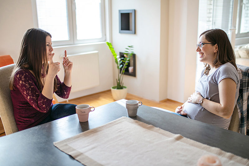 Women seated at table conversing in sign. One is pregnant and holding her stomach.