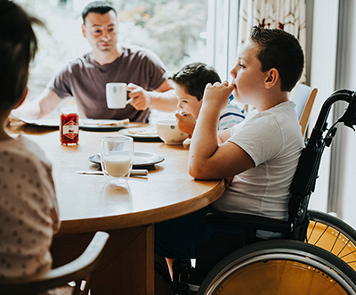 A boy sitting in a wheelchair at a table, eating a meal with his family.