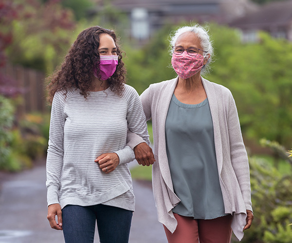 Two women wearing protective face masks link arms and walk together.