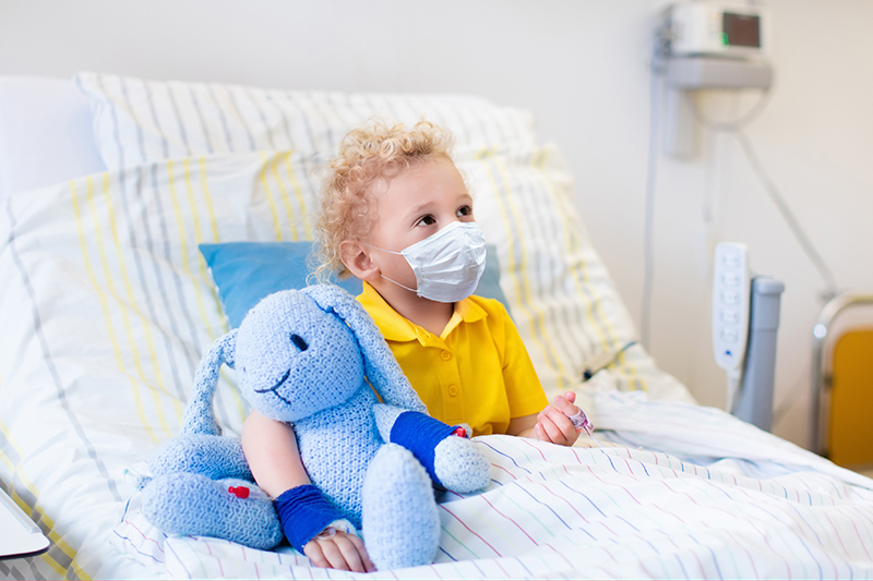 Masked child sitting in hospital bed holding a stuffed toy.