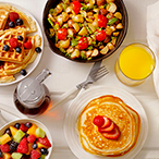 A white table with an assortment of beverages and plates of breakfast food, including waffles, pancakes, fruit, and croissants.