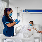 A girl is in a hospital bed flanked by a healthcare provider, who is filling out a chart, and her father, who is sitting down and holding her hand.