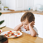 Child sitting at table, looking sadly at plate of food.