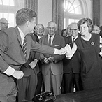 A black and white photo of President John F. Kennedy reaching for a pen in a crowded room.