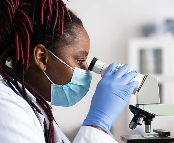 A masked researcher looks through a microscope in a laboratory.