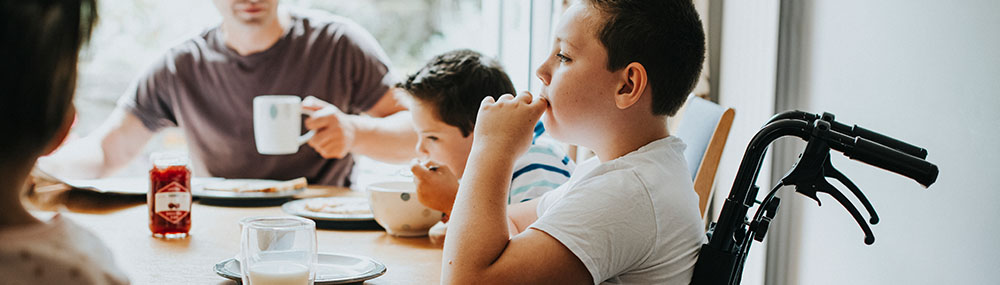 Young child sitting in a wheelchair, having a meal with his family at a kitchen table.