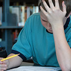 A teenage boy sits at a table in front of a bookshelf. He is hunched over a piece of paper. He holds a pencil in his right hand, and his left hand presses against his forehead.