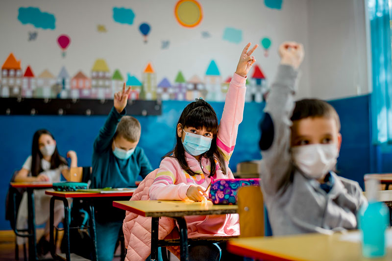 Children in classroom seated at their desks, raising their hands.