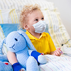 A boy sits up in a hospital bed. He’s wearing a mask and holding onto a blue stuffed bunny.