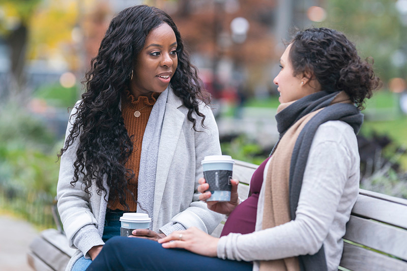 A Black woman and her pregnant White friend sit on a park bench having coffee and talking.