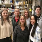 Six people stand in a laboratory in front of shelves that contain various bottles and boxes.