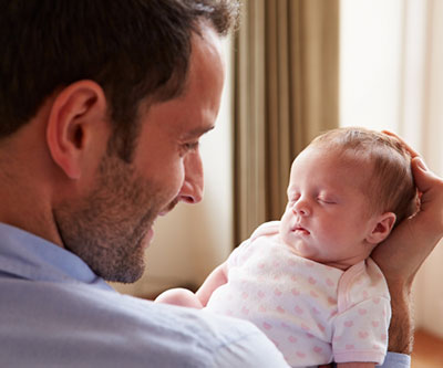 A smiling father gently cradles a sleeping newborn baby in his hands. 