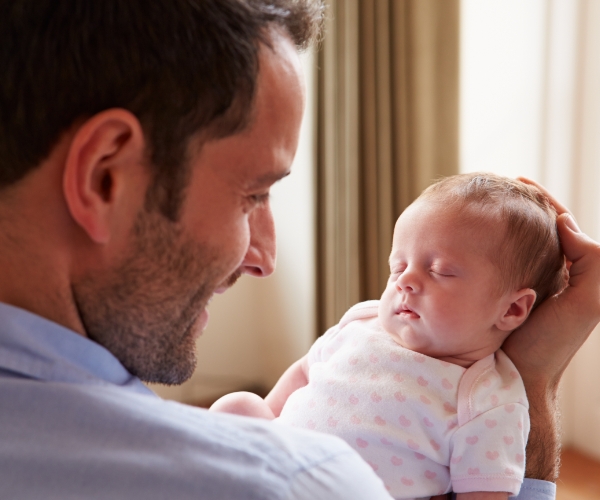 A smiling father gently cradles a sleeping newborn baby in his hands.