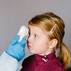 Healthcare provider wearing mask and gloves scans a child’s forehead with a digital thermometer.