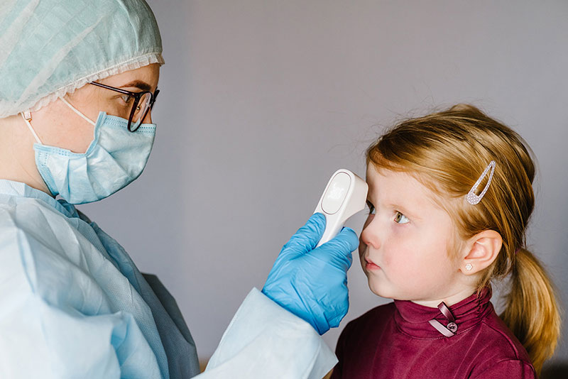 Healthcare provider wearing mask and gloves scans a child’s forehead with a digital thermometer.