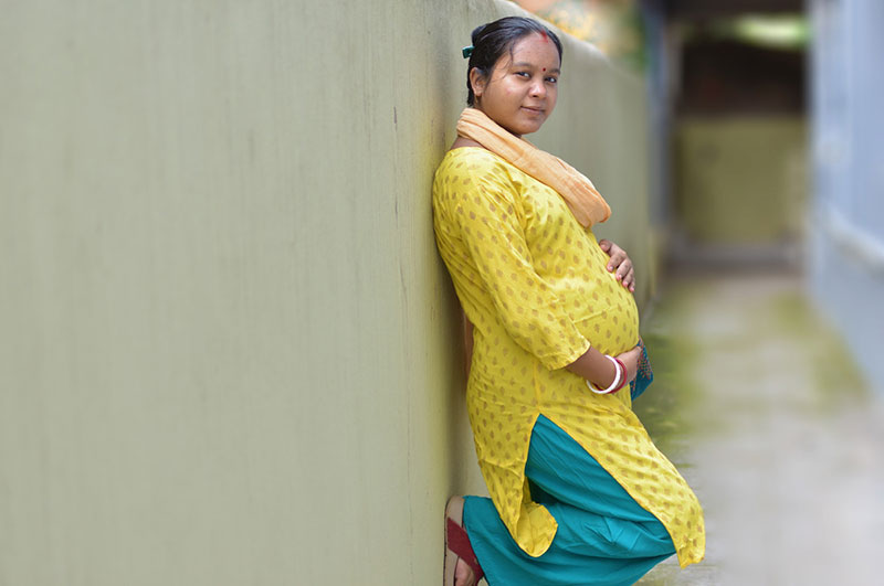 A pregnant woman wearing a yellow tunic, teal pants, and red sandals stands in an alley with her hands on her belly.