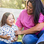 A little girl with Down syndrome sits on the grass with her adult female caretaker. Both are laughing. Trees and a stone wall are visible in the background.