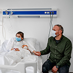 Child in hospital bed, parent seated at right. All are masked.