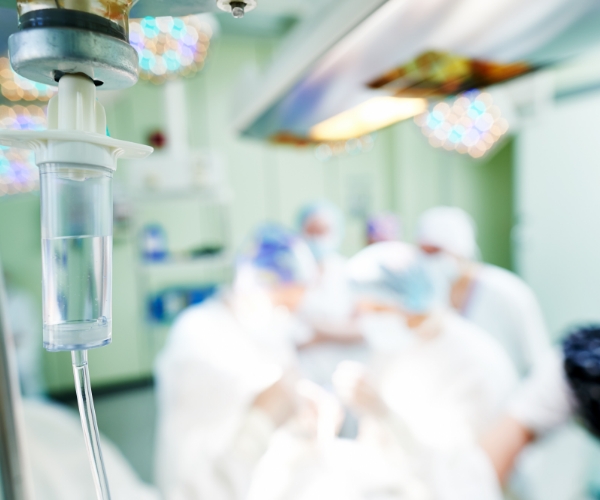 An IV drip chamber appears in sharp focus in the foreground, with blurred medical staff and patient in the background. 