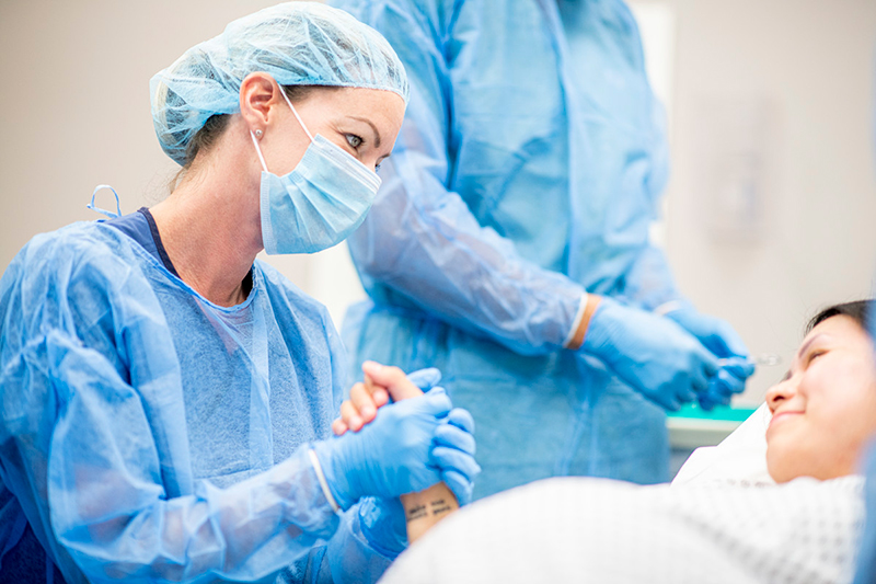 Health care worker in surgical garb holds hand of pregnant person lying on gurney. Upper torso of health care worker in surgical garb visible in background.