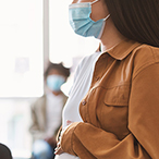 Masked medical professional writing on a desk top while a masked pregnant person looks on.