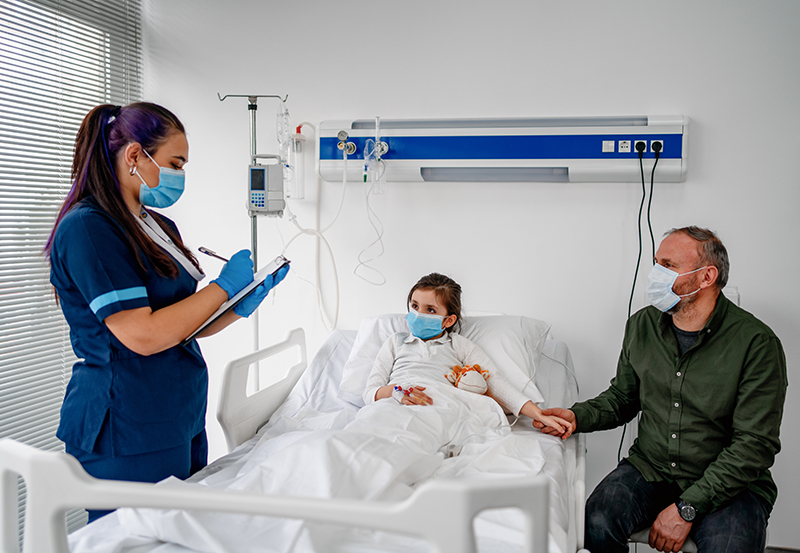 Child lying in hospital bed, concerned parent at bedside. Health care worker writing on clipboard. All are masked.