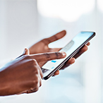 Close-up photo of the hands of a Black female health care worker typing on a cell phone in front of a blurred background. A white coat and stethoscope are partially visible on the left of the image.