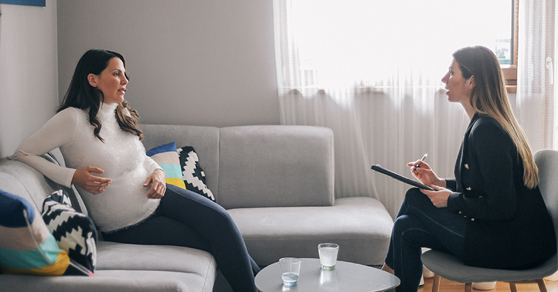 Pregnant person, seated on couch, talking to mental health care professional with note pad and pen.