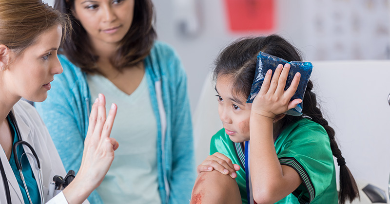 Health care worker holds up three fingers before child holding ice pack to head while parent looks on.