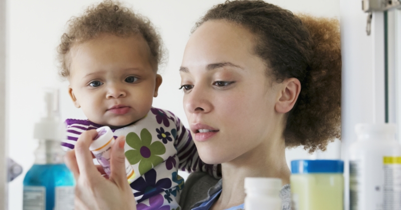 A woman holding a baby reads the label on a bottle of prescription medicine. Other medicine cabinet items appear blurred in the foreground.