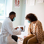 Masked health care provider in white lab coat writes on clipboard while masked patient seated on examination table looks on.