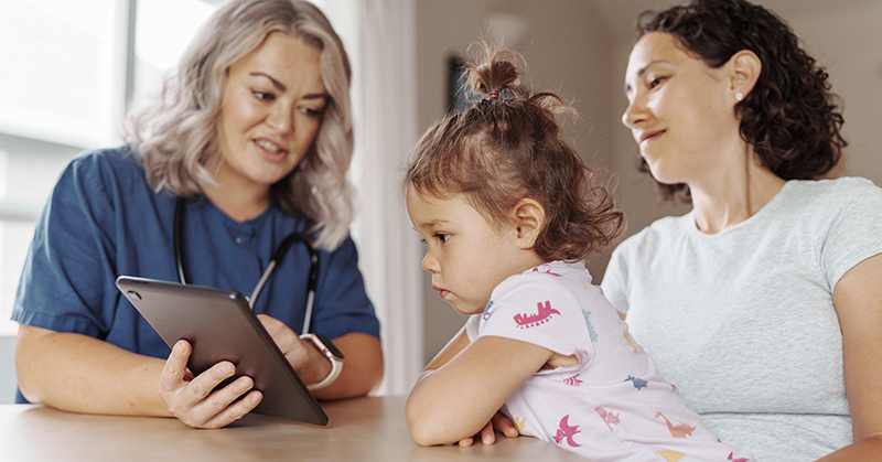 A toddler sitting on her mother’s lap gazes at a tablet device held by a health care provider.