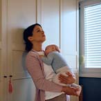 Parent leaning against a wall, gazing at ceiling while holding infant.