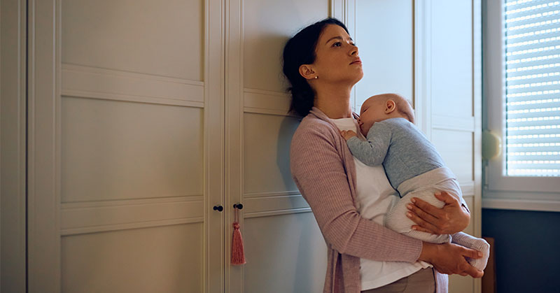 Parent leaning against a wall, gazing at ceiling while holding infant.